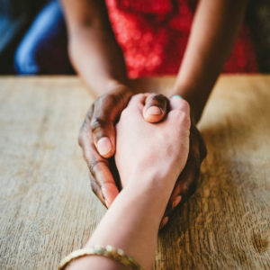 Two people clasp hands in prayer over a wooden tabletop