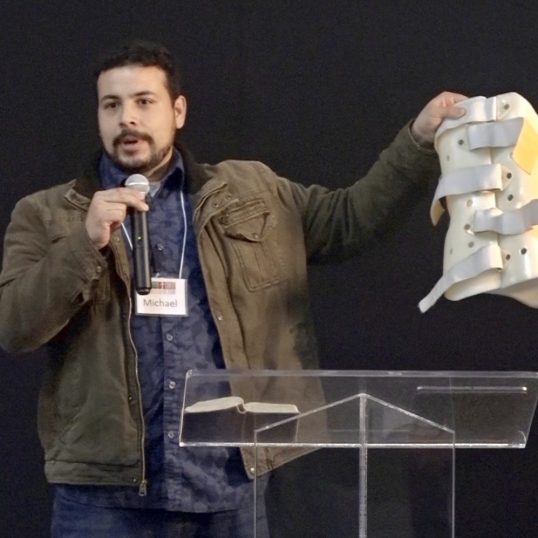 A man holding a microphone and speaking at a podium, holds out the back-brace he no longer needs