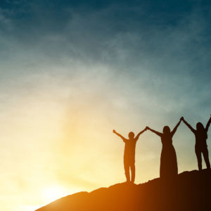 A trio is silhoutted at sunset on a rocky hill, their arms raised in a v, holding hands