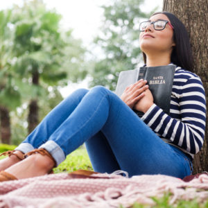 A young woman, sitting against a tree, holds her bible to her chest with both hands, her eyes closed