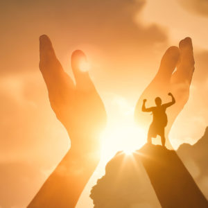 Double exposure of raised hands and a silhoutted person with arms raised against a glowing sunrise