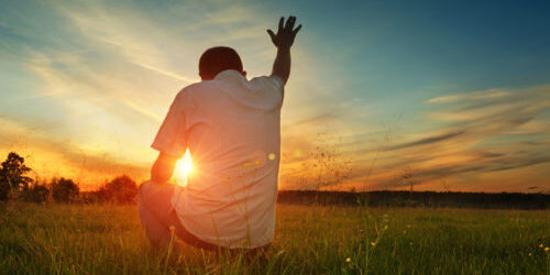 A man holds his hand up to the sky and kneels in prayer as the sun sets