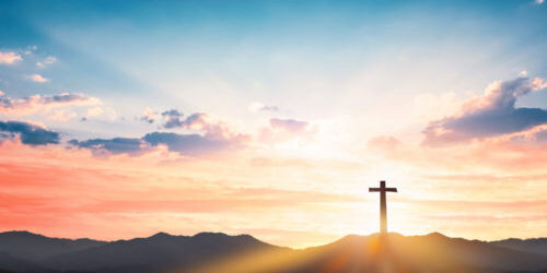 A cross sits high on a mountaintop, backlit by a beautiful sunrise