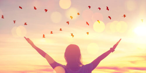 Woman stands at sunrise with her arms raised in a v, birds fly by and glowing light surrounds her