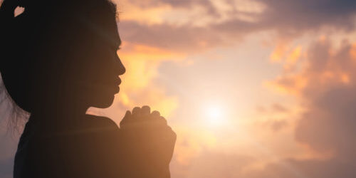 A woman's praying profile is silhoetted by the setting sun peaking through the clouds