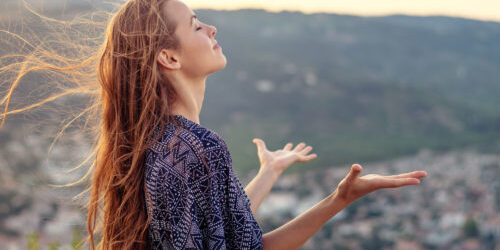 A blissful young woman with long hair stands in prayer on a hilltop at dusj